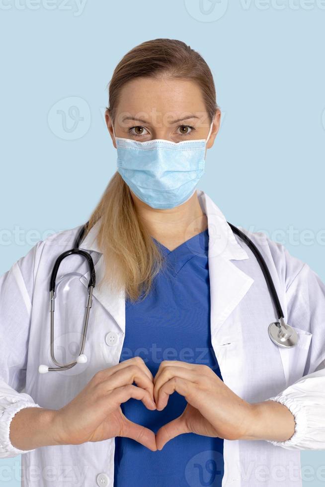 Portrait of young female doctor with medical mask, showing heart shape with hands. Love symbol. Medicine, healthcare during Covid-19. Doctor with white coat, stethoscope over neck, ready to help. photo