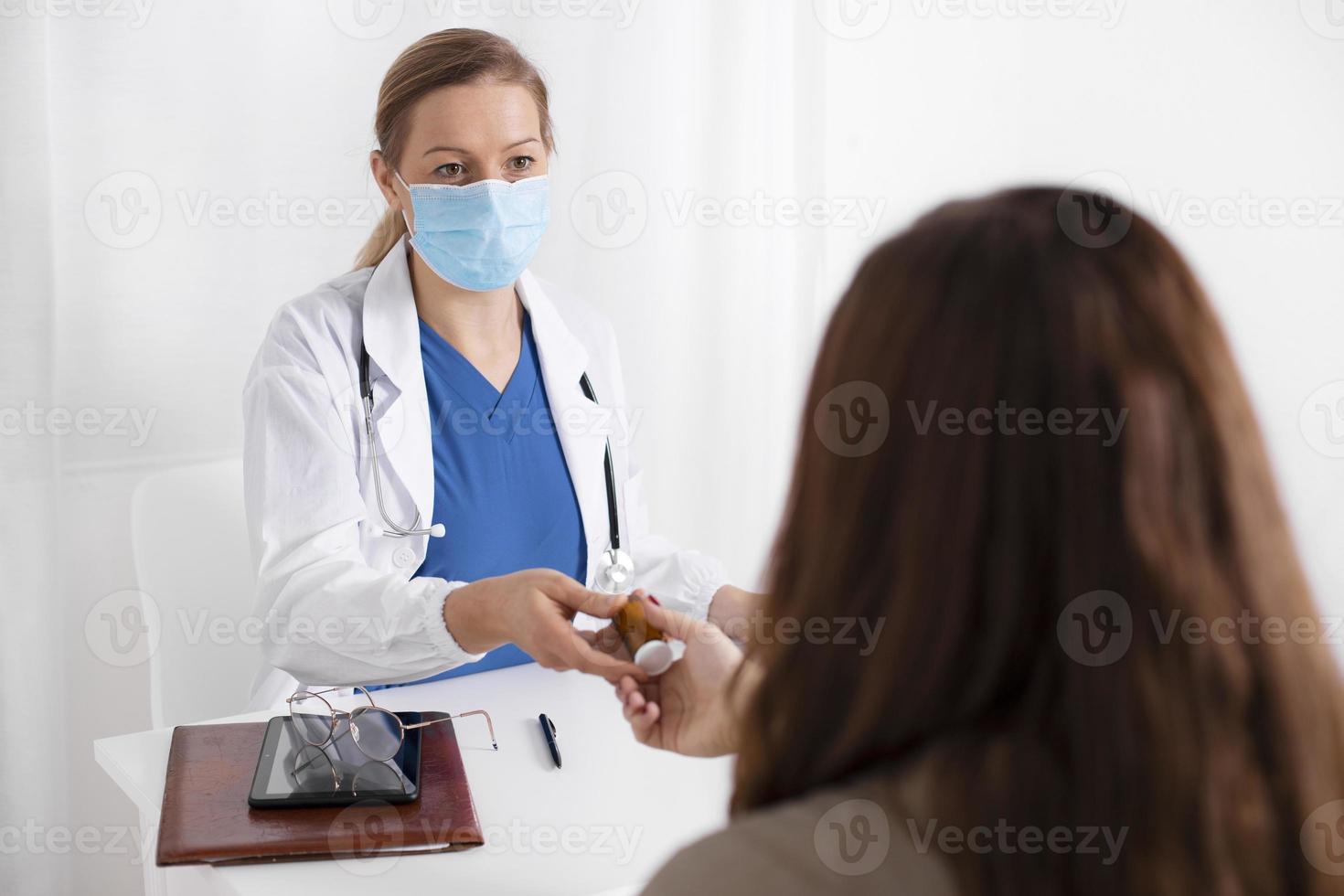 Young brunette woman having consultation at doctor office during coronavirus and flu outbreak. Female doctor wearing facemask, giving to patient jar with pills. Virus protection. Covid 19. photo