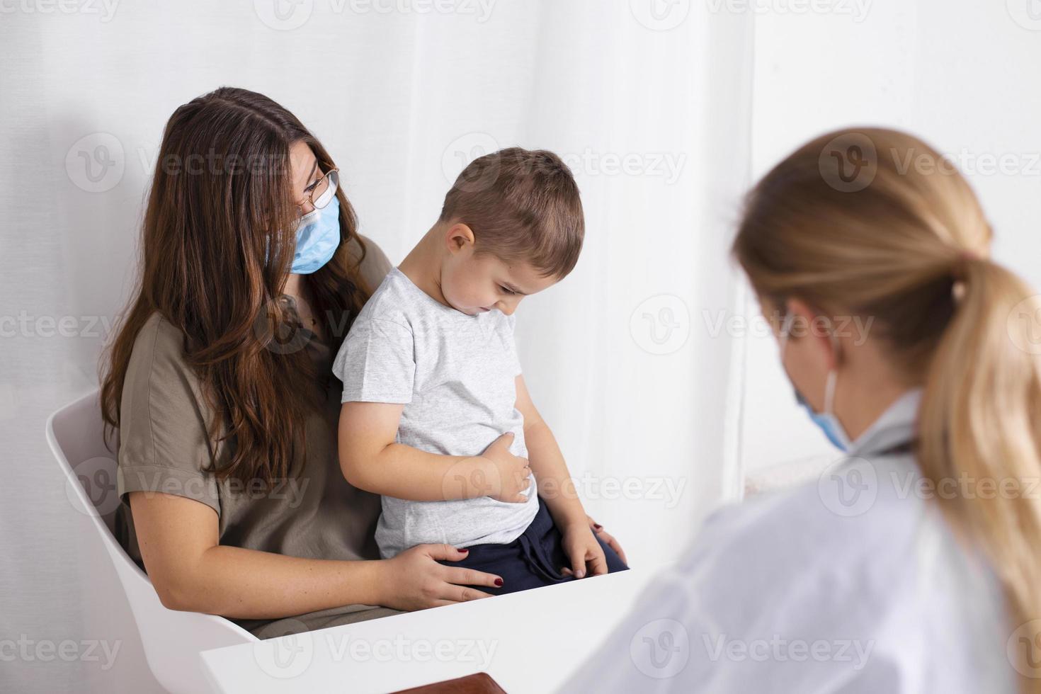 Young brunette woman with little boy having consultation at pediatrician office. Child has abdominal pain. Doctor, child and mother wearing facemasks during coronavirus and flu outbreak. Covid 19. photo