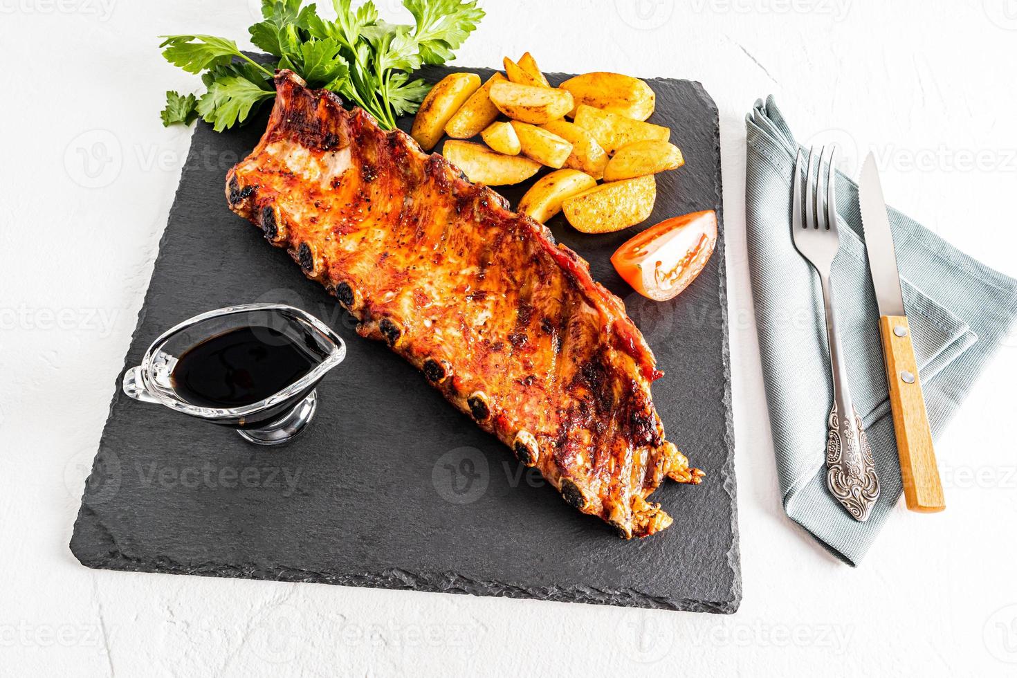 grilled pork ribs on an slate dark board with slices of potatoes, tomatoes, sauce. gourmet lunch. delicious food. white background. photo