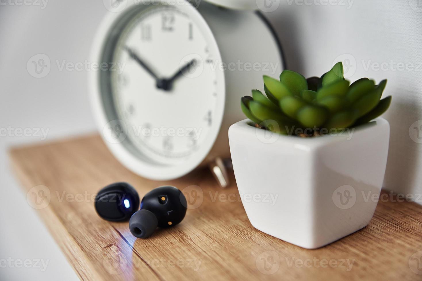 Wireless earphones on shelf with alarm clock and house plant photo