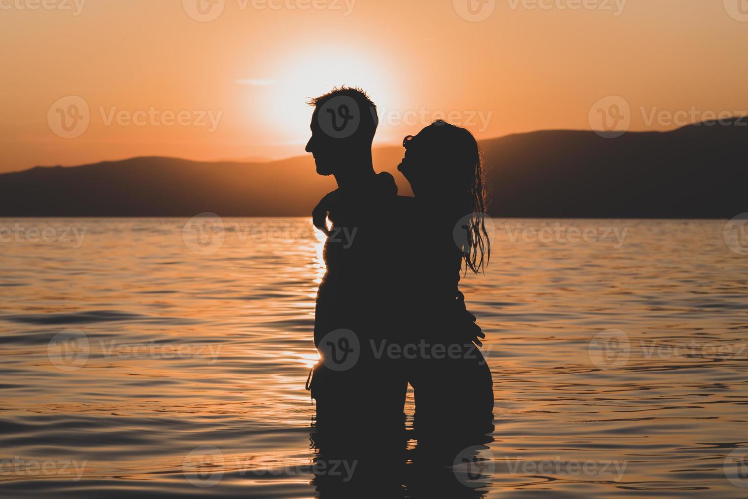 siluetas enamoradas amantes de la pareja romántica abrazándose, besándose, tocándose, contacto visual al atardecer, amanecer en el fondo del mar, el sol, las nubes en colores rojo fuego, naranja foto