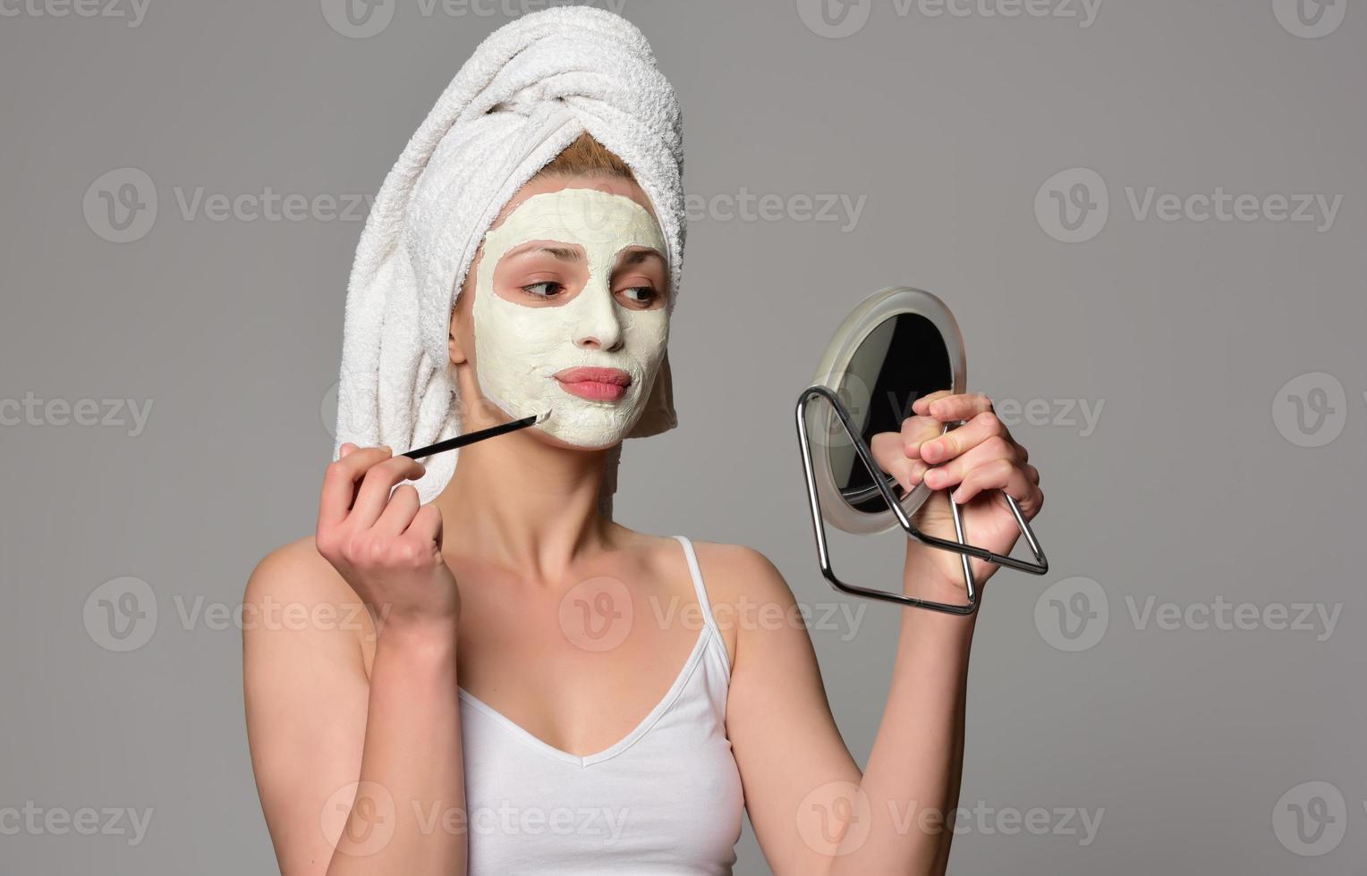 Beautiful female model with towel on her hear and white facial cosmetic mask on her face. Beauty cosmetic concept. Isolated on gray background. photo