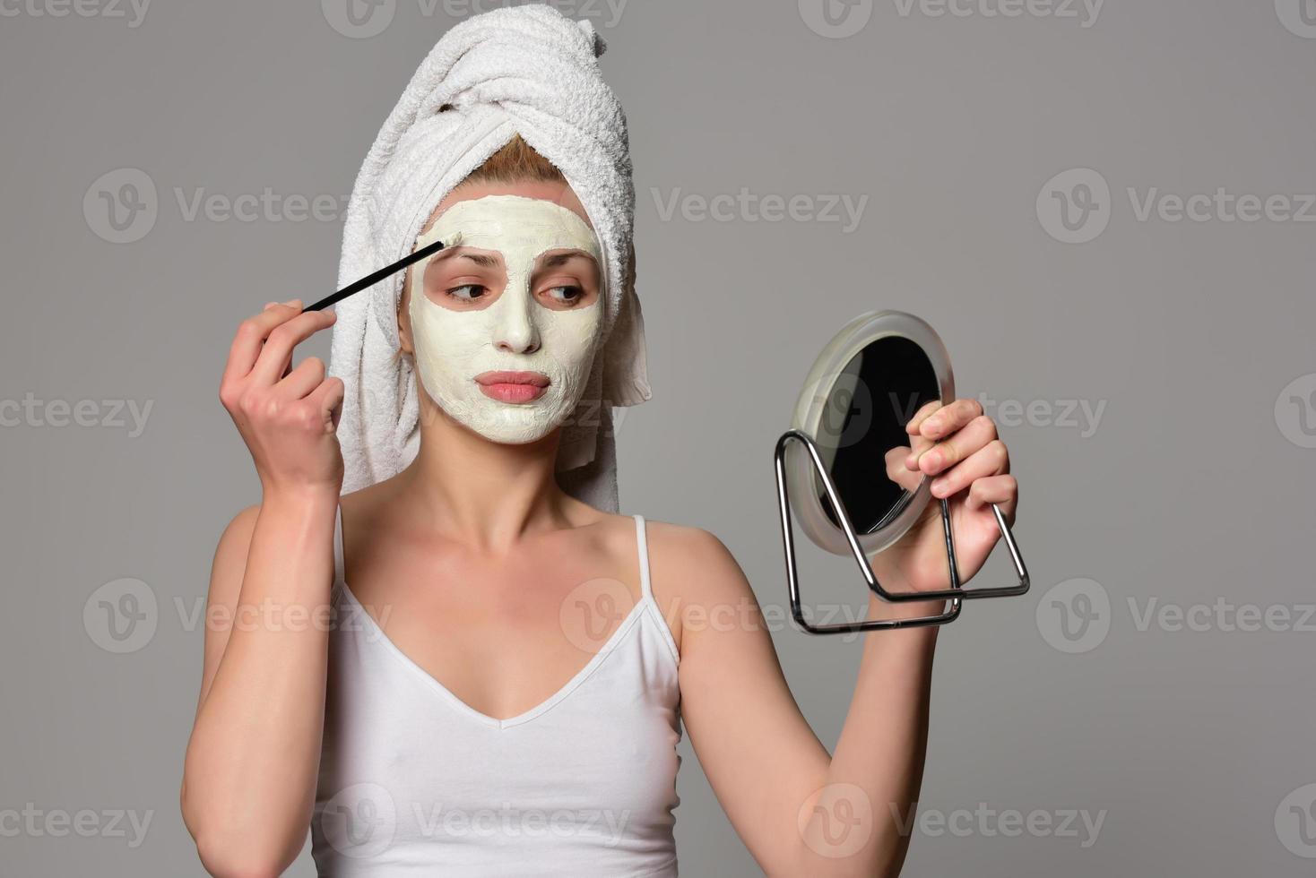 Beautiful female model with towel on her hear and white facial cosmetic mask on her face. Beauty cosmetic concept. Isolated on gray background. photo