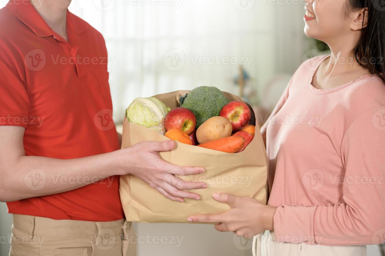 Asian young man delivery in red uniform holding healthy fruit vegetables in grocery bag send to customer in kitchen background photo