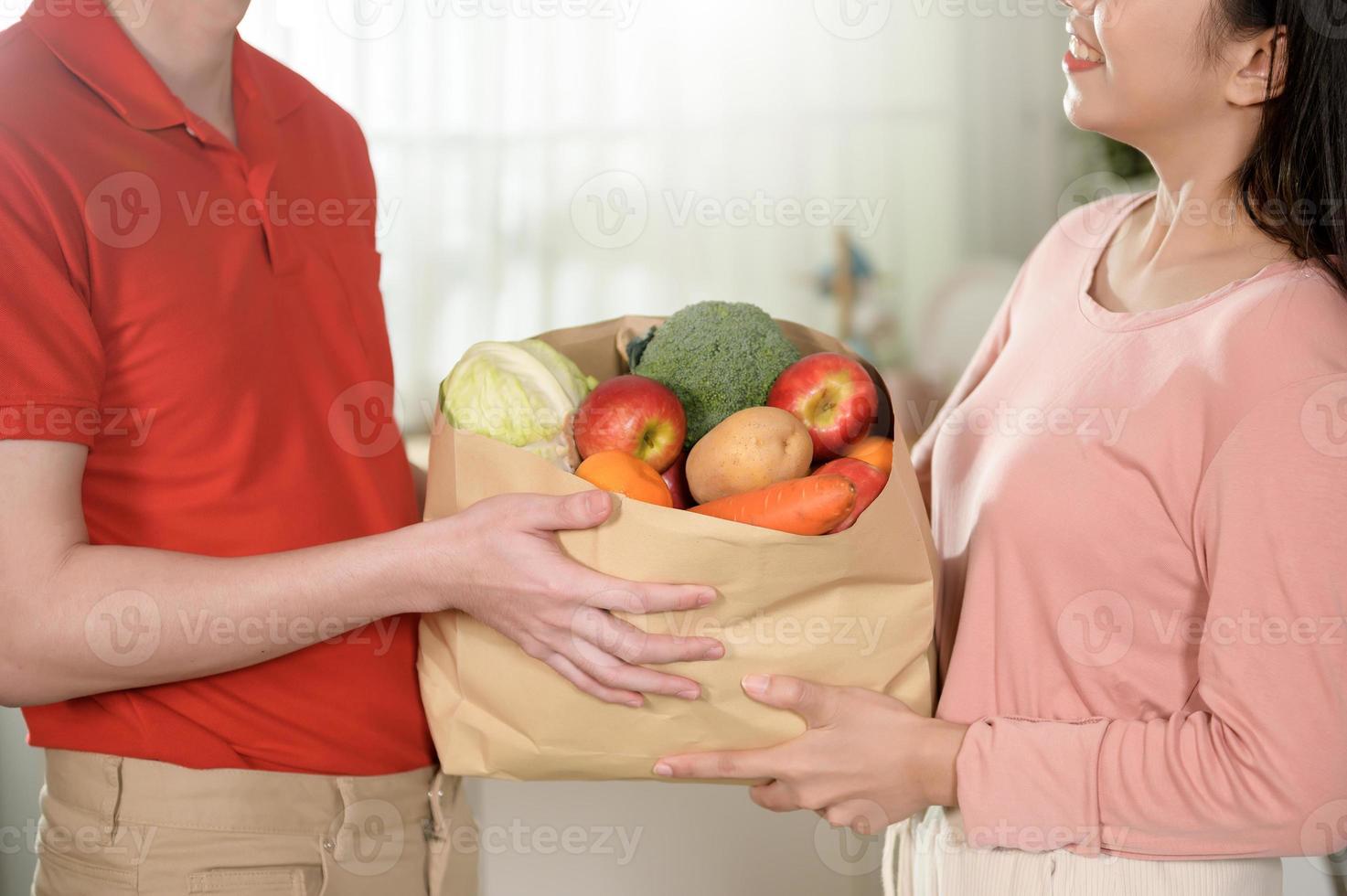 entrega de un joven asiático con uniforme rojo sosteniendo verduras de frutas saludables en una bolsa de supermercado enviada al cliente en el fondo de la cocina foto