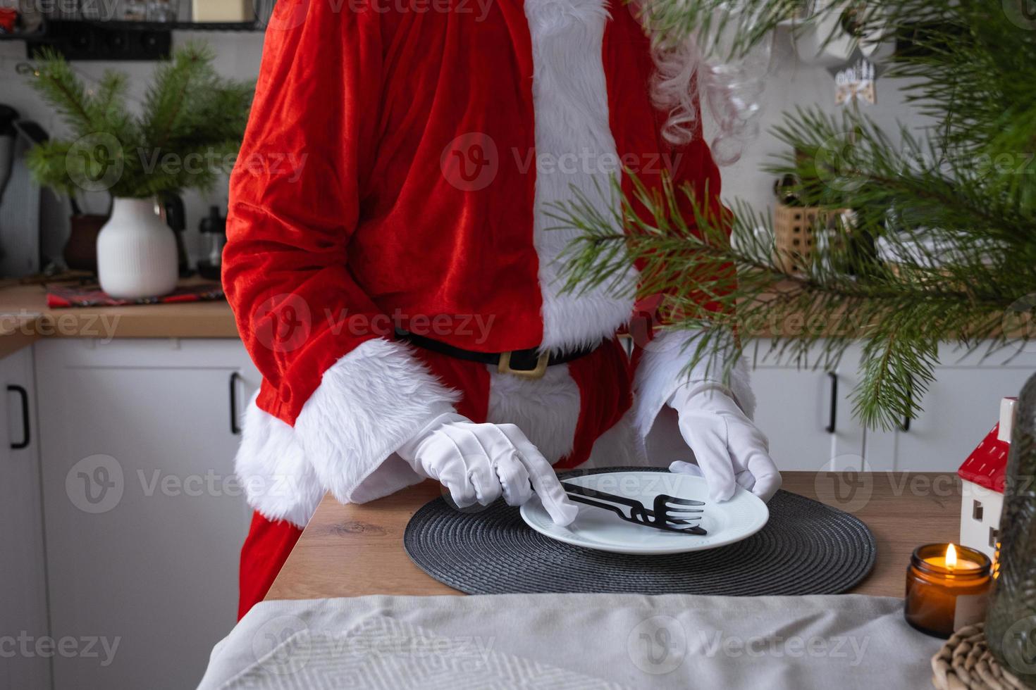 santa claus pone la mesa en casa en la cocina blanca, pone un plato, pone cubiertos, tenedor y cuchillo. preparándose para la celebración de la navidad, poniendo la mesa para los invitados foto