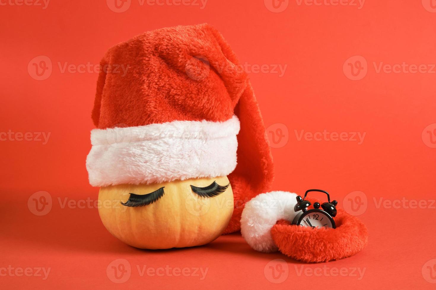 pumpkin in santa hat and false eyelashes and black little alarm clock on red background copy space photo