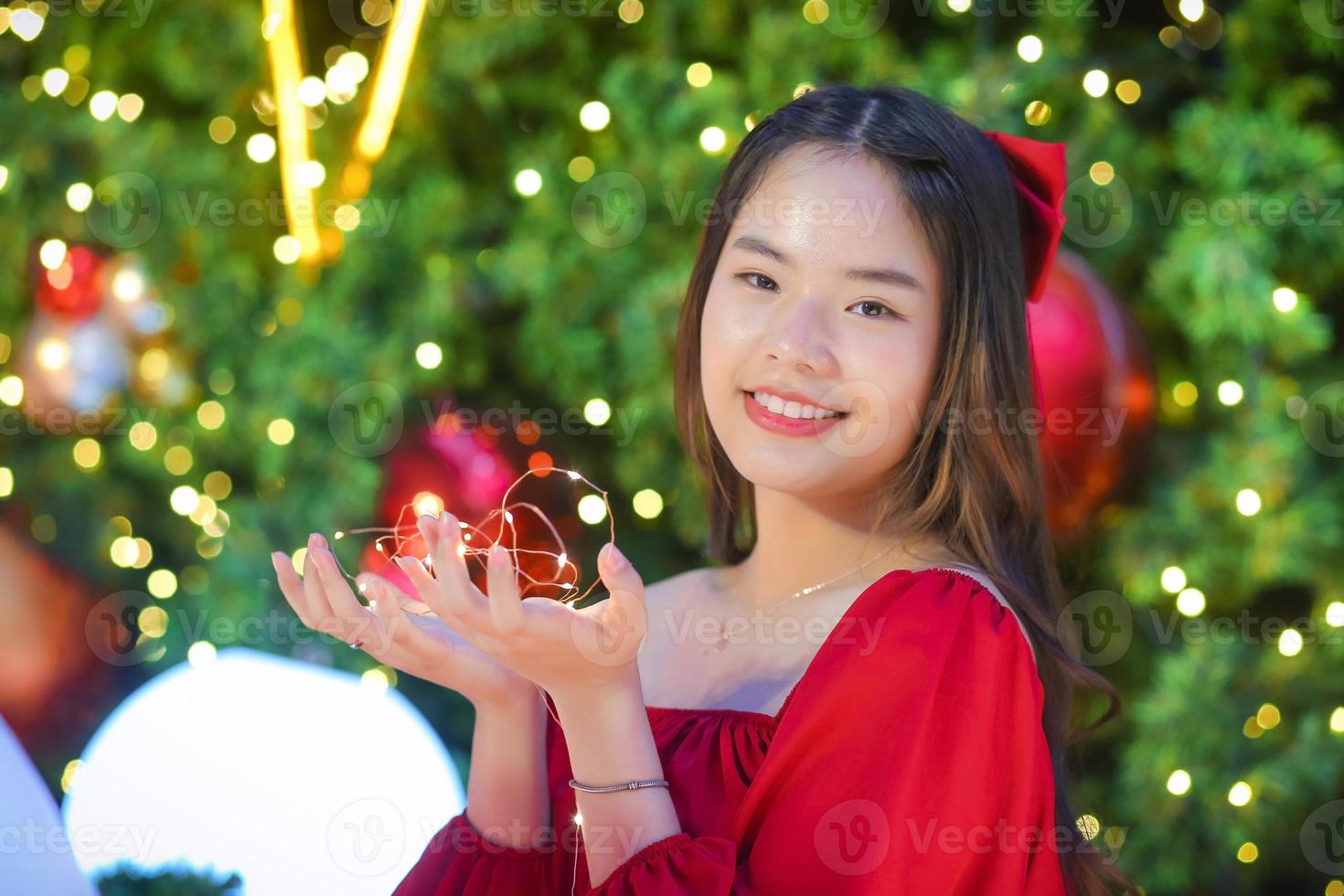 bella mujer asiática vestida de rojo se pone de pie felizmente. en su mano sostenía una luz frente al árbol de navidad. con bokeh como fondo en el tema de las celebraciones de navidad y año nuevo foto