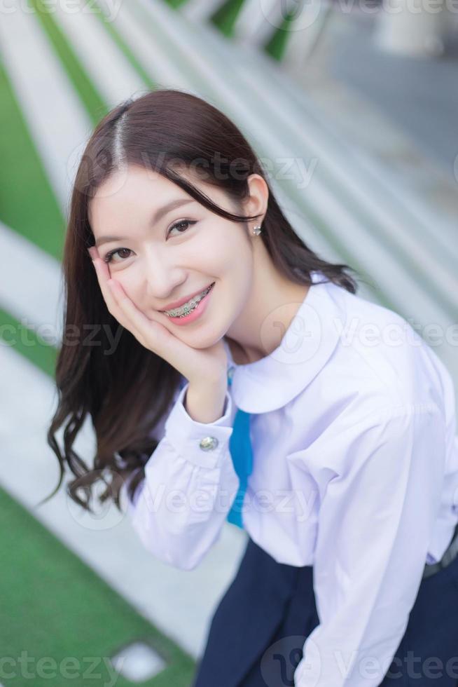 Beautiful high school Asian student girl in the school uniform stands and smiles happily with braces on her teeth while she put her hand on face confidently with the building as a background. photo
