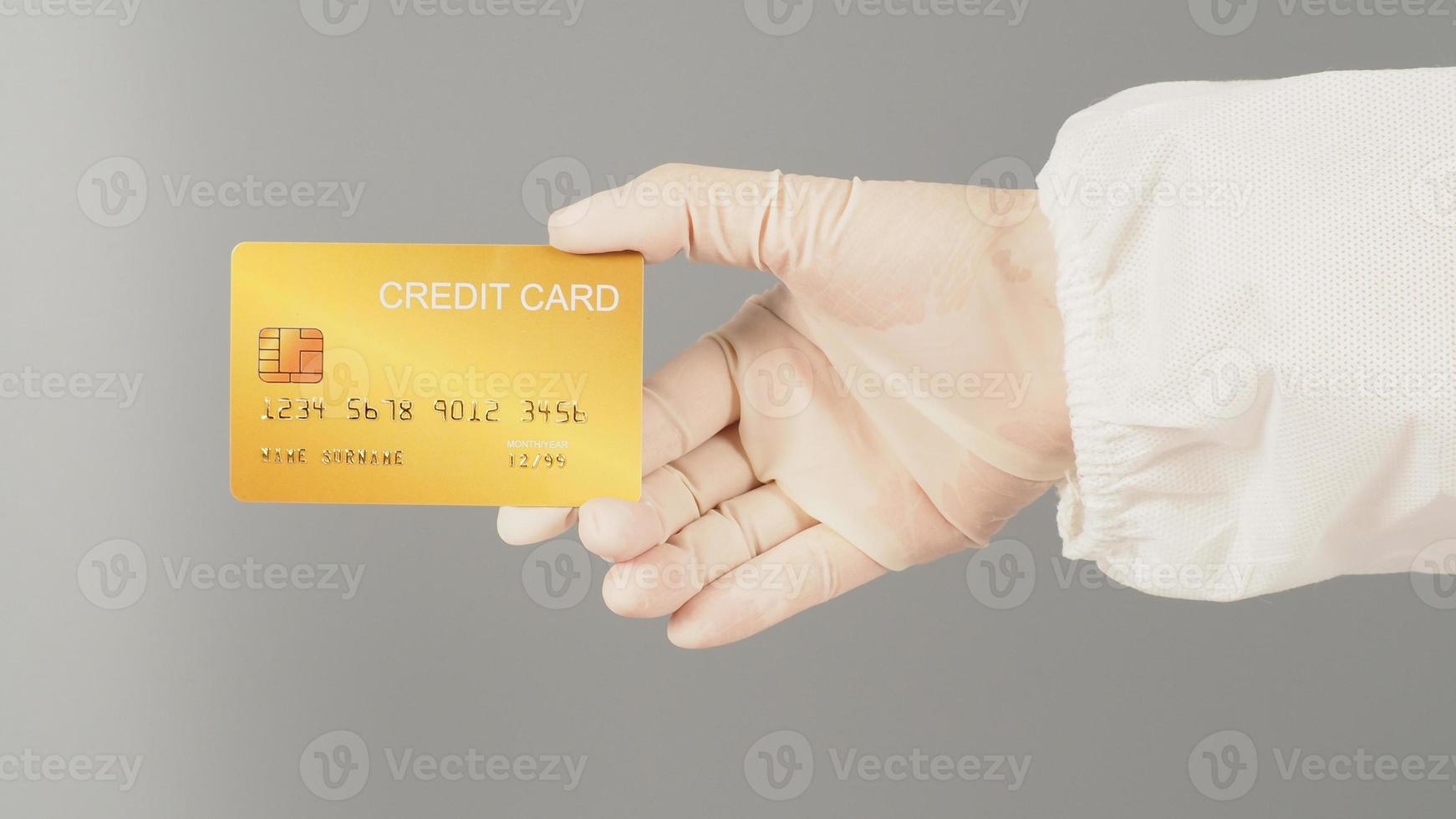 la mano sostiene una tarjeta de crédito dorada sobre fondo gris. la mano lleva un traje ppe y un guante médico blanco. foto