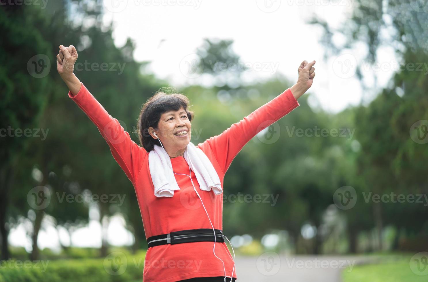 Athletic asian senior woman 60s smiling, hands up and happy cheerful with success and jogging. Senior asian woman running at park. Female runner listening to music with earphones while jogging. photo
