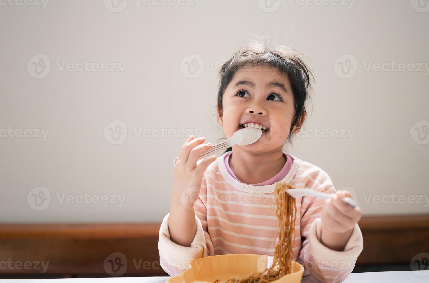 la niña asiática disfruta feliz usando cubiertos, cuchara y tenedor comiendo deliciosos fideos en la cocina en la mesa del comedor. una niña asiática feliz practica comer sola en la mesa del comedor. concepto de comida para bebés foto