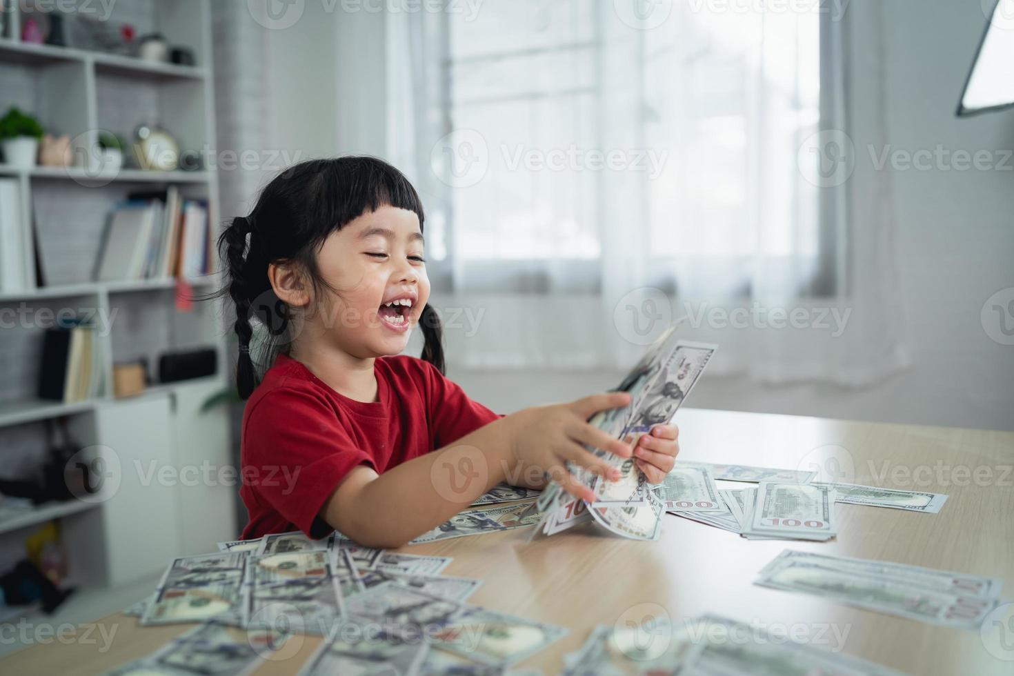 Asian baby girl wearing a red t-shirt holding dollar bill on wood table desk in living room at home. Saving investment wealth, growth stock, invest interest, fund, saving money for the future concept. photo