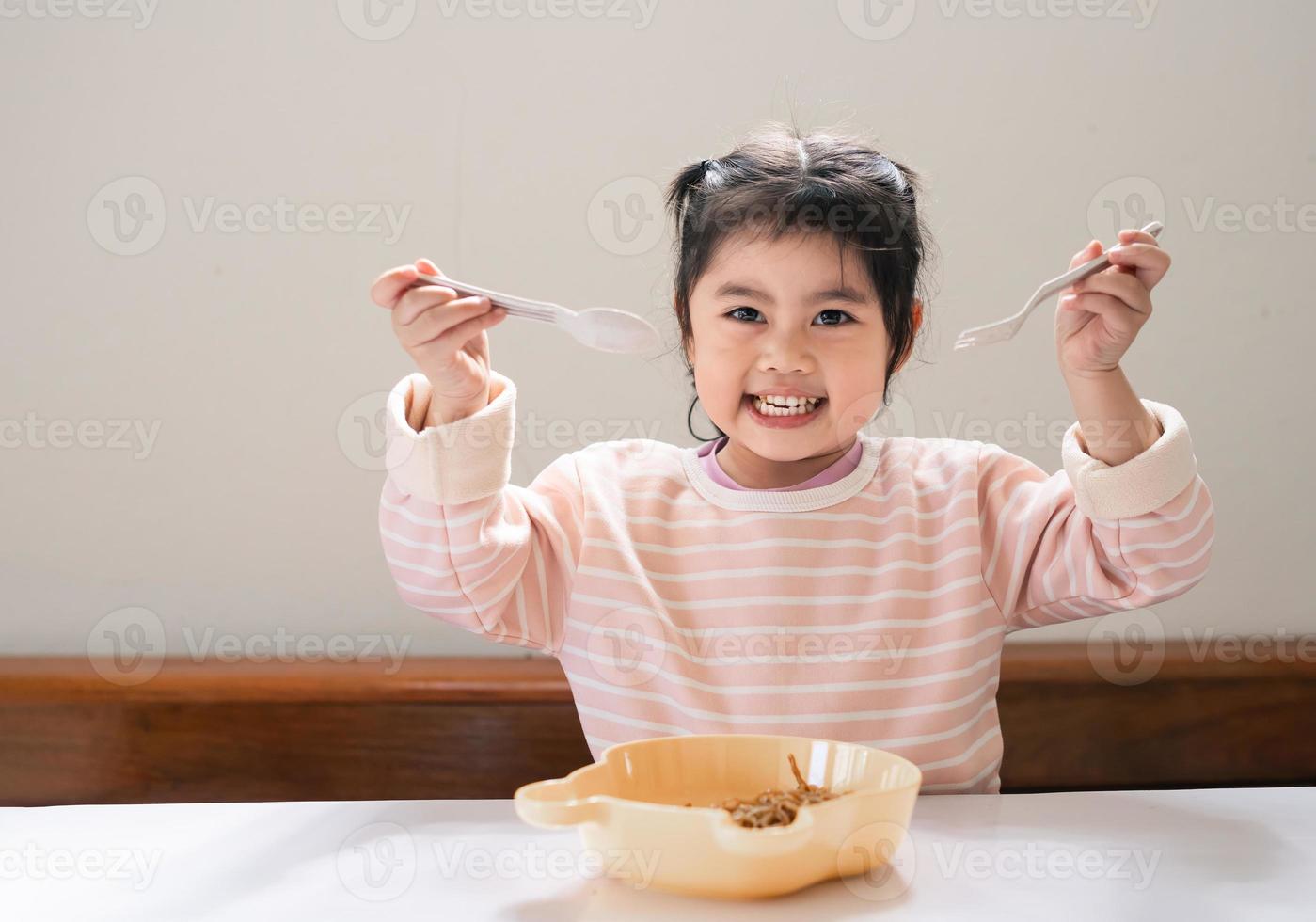 Asian baby girl enjoy happy using cutlery spoon and fork eating delicious noodle in kitchen on dining table. Happy asian baby girl practice eating by her self on dining table. Baby food concept photo