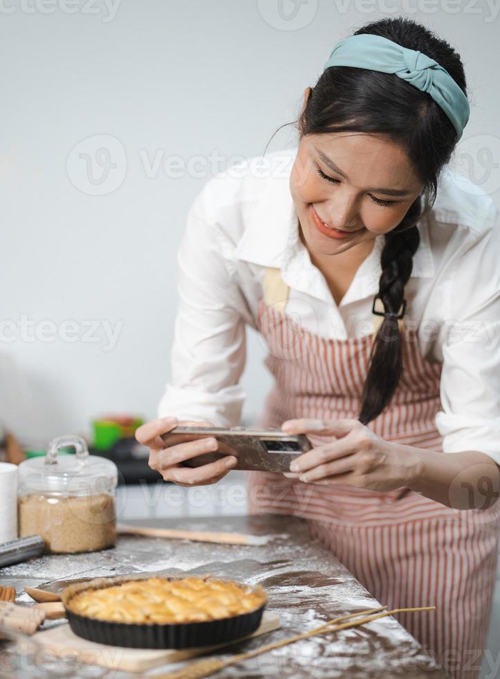 mujer joven usa delantal tomando una foto selfie con pastel casero en la cocina. retrato de una hermosa mujer asiática horneando postres y divirtiéndose tomando fotos con un smartphone para redes sociales en línea. cocina casera.