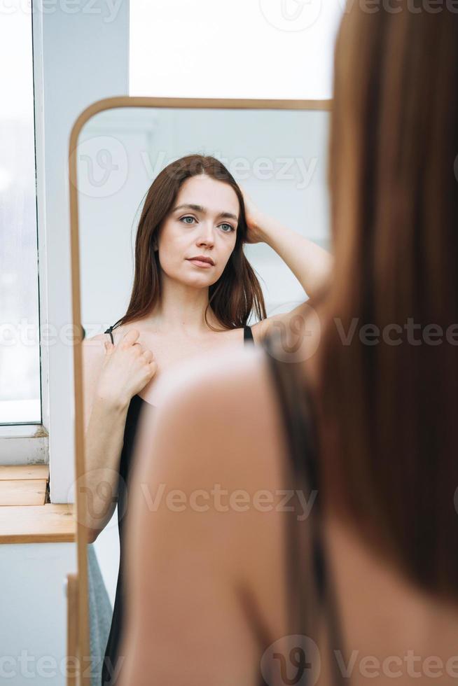 Young woman with long dark hair on evening elegant black dress with open back near mirror in white interior photo