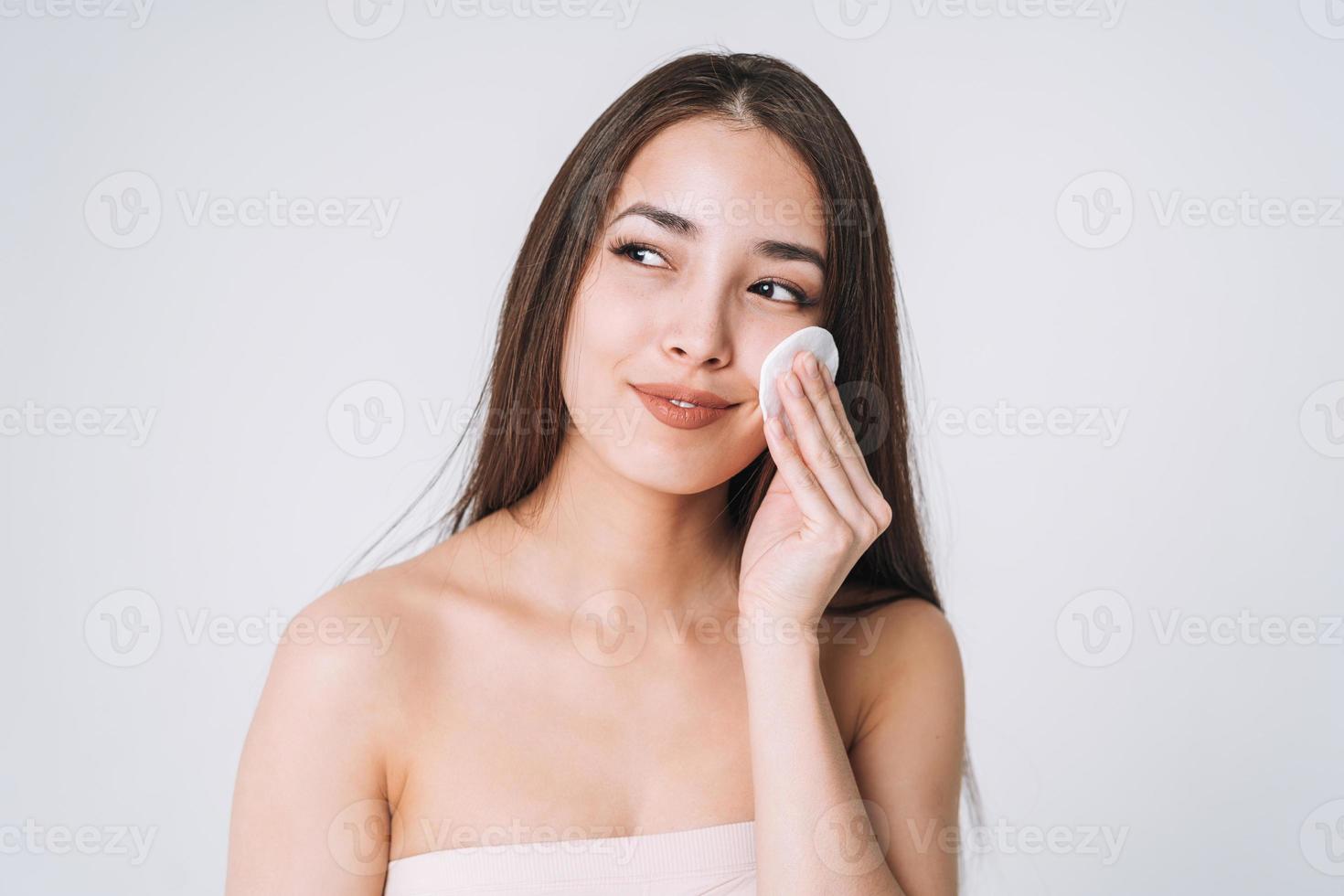 Beauty portrait of happy smiling asian woman with dark long hair with cotton pad on clean fresh skin face and hands on white background isolated, skin cleansing and removing makeup photo