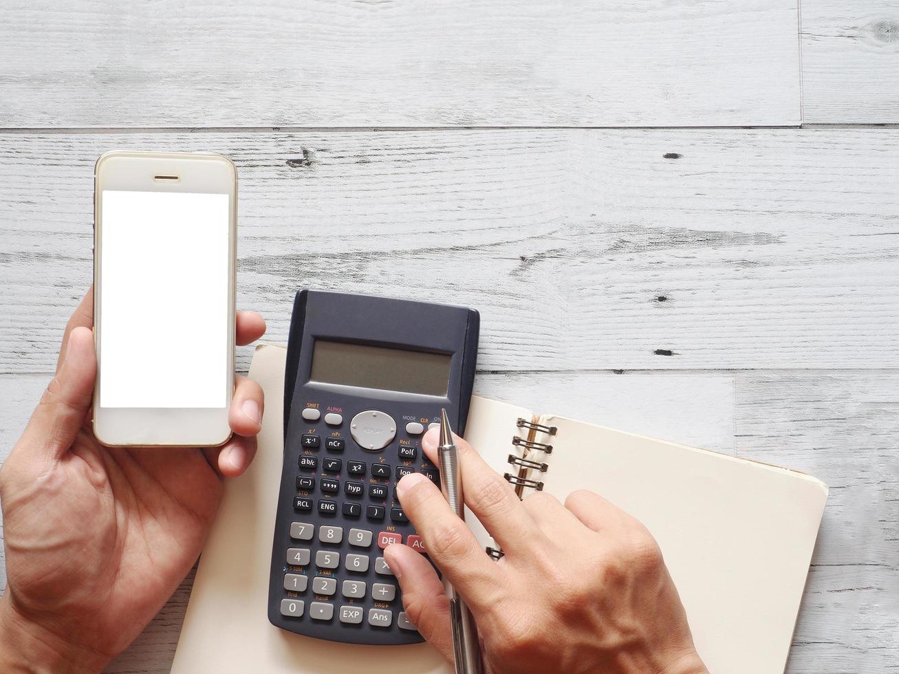 Hand holding mobile phone white screen and using calculator with cactus and silver pen on white wood table nature shadow and sunlight top view and space business concept photo