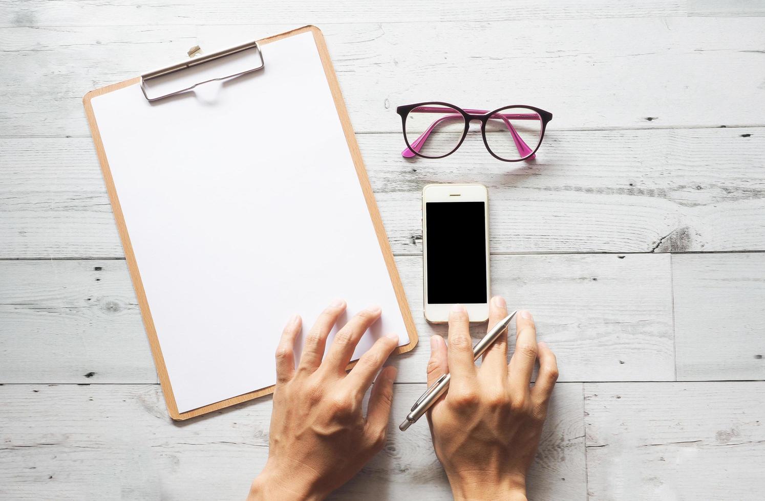 Hand with silver pen on clipboard and glasses and using smartphone black display on white wood table nature shadow top view photo