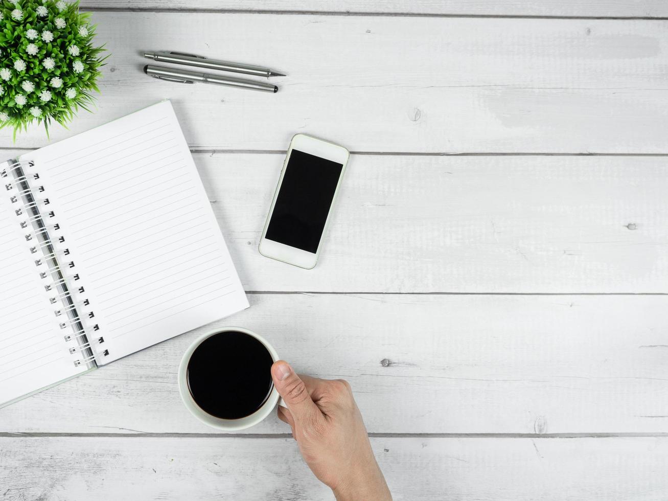 espacio de trabajo en el espacio de copia de la vista superior del escritorio de madera blanca, página en blanco del cuaderno con pantalla negra del teléfono inteligente y bolígrafo plateado con taza de café de mano foto