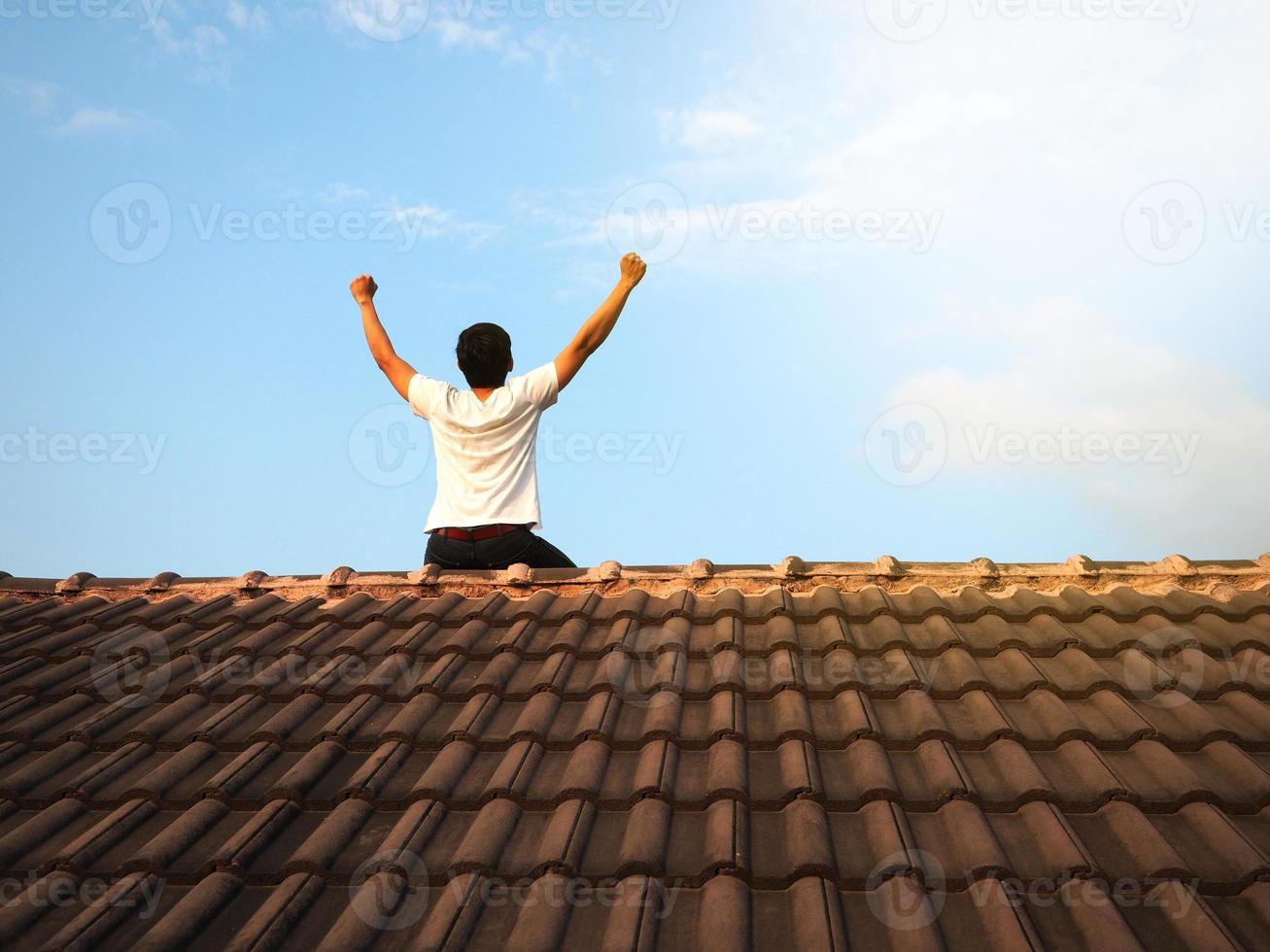 Man sit on the roof with blue sky background success concept,Fighting man sit turn back on black roof and show arm up photo