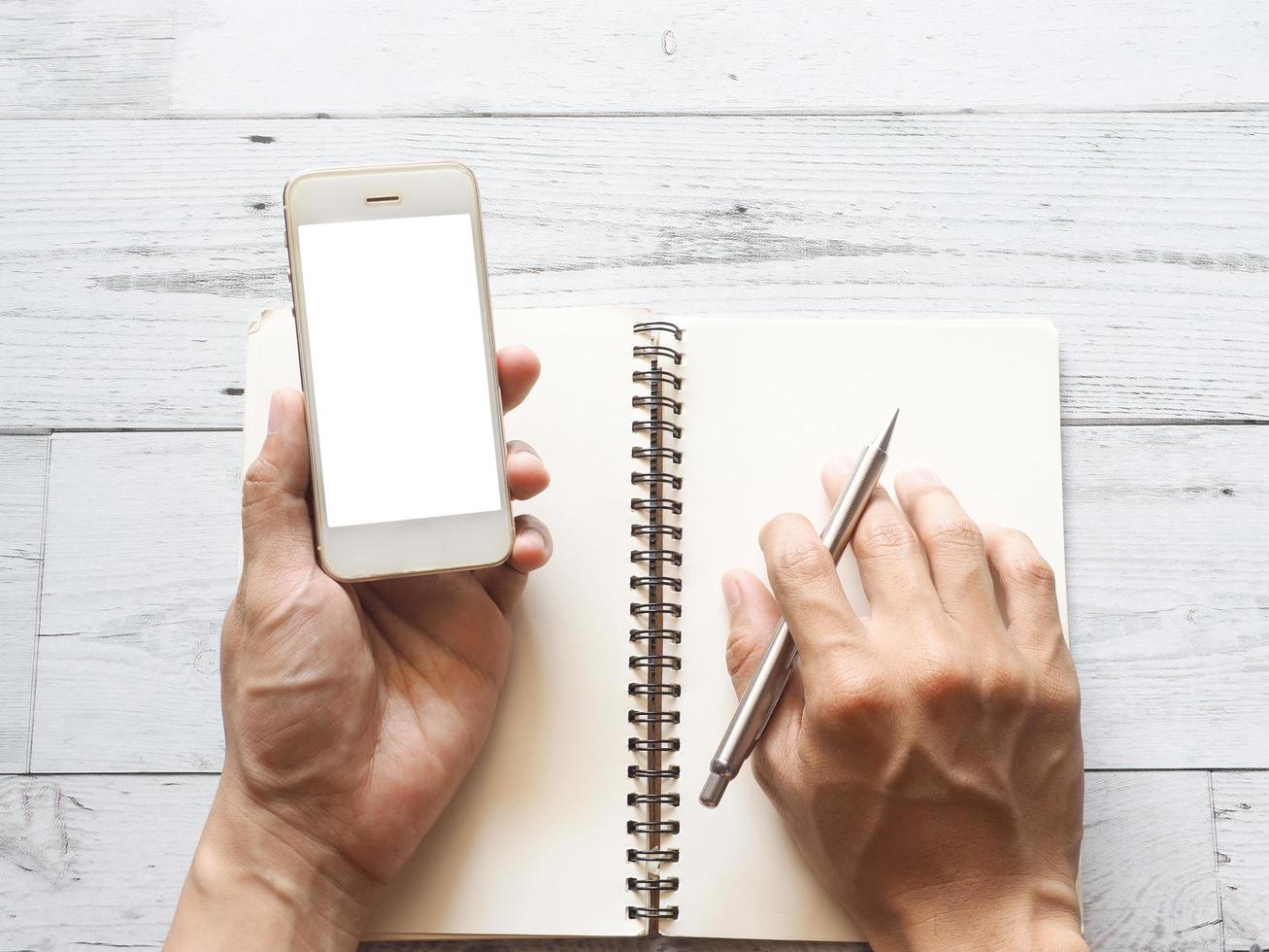 Hand holding smartphone white display and writing on blank page notebook by silver pen on white wooden table top view space nature shadow photo