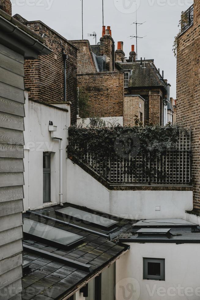 Side street of Notting Hill in London with typical brick walls and chimneys on the roofs of england houses with wooden fence covered with green bush which separate the backyard from the white building photo