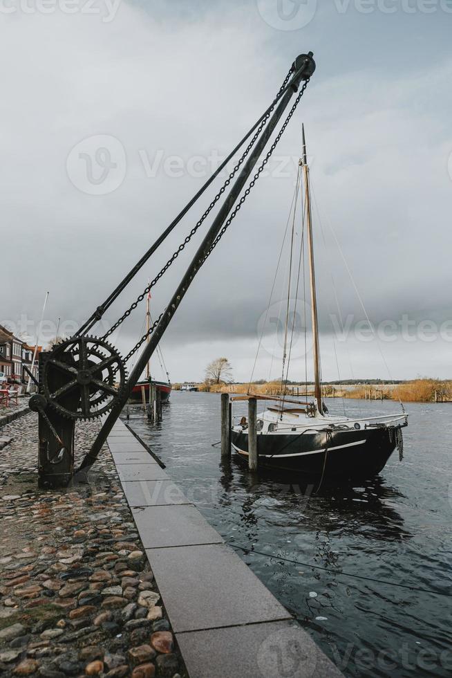 viejo velero en el río inundado en el puerto de la ciudad de ribe en dinamarca con la vieja grúa del puerto en frente durante el día nublado de otoño en orientación vertical foto