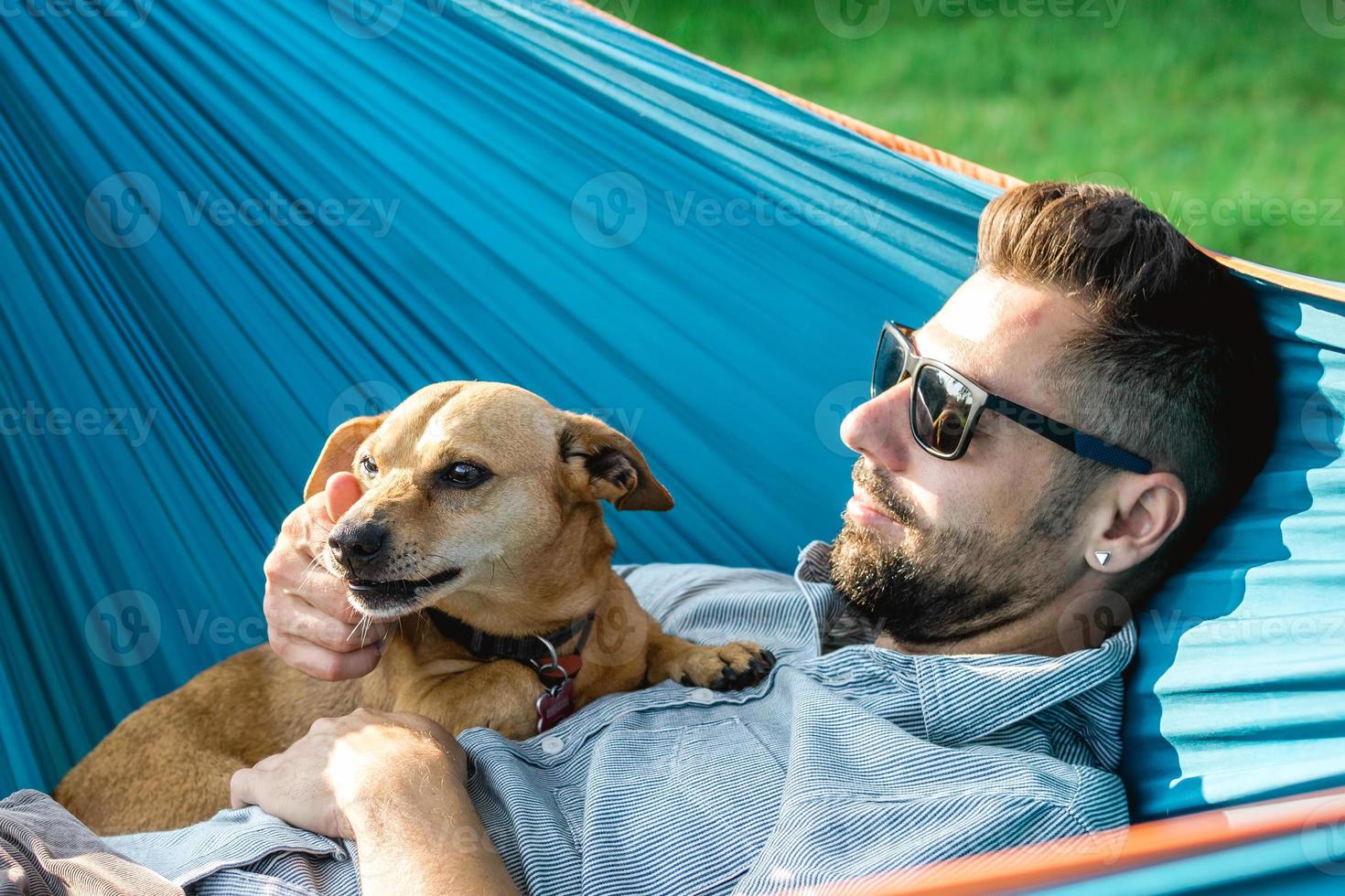 Handsome European man in sunglasses is resting in hammock with his cute little dog. photo