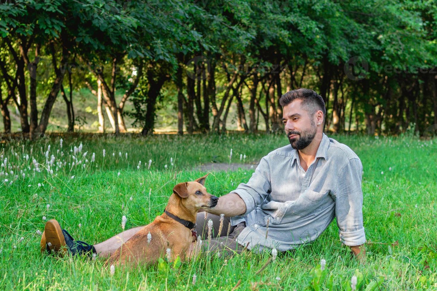 hombre guapo con gafas de sol sentado en el césped con su perro en el parque. concepto de relación humana y mascota. foto