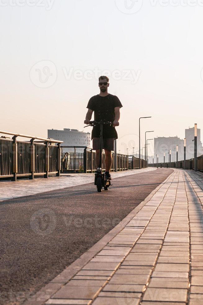 Attractive European man is riding on electric scooter on empty street of city at sunny day. photo