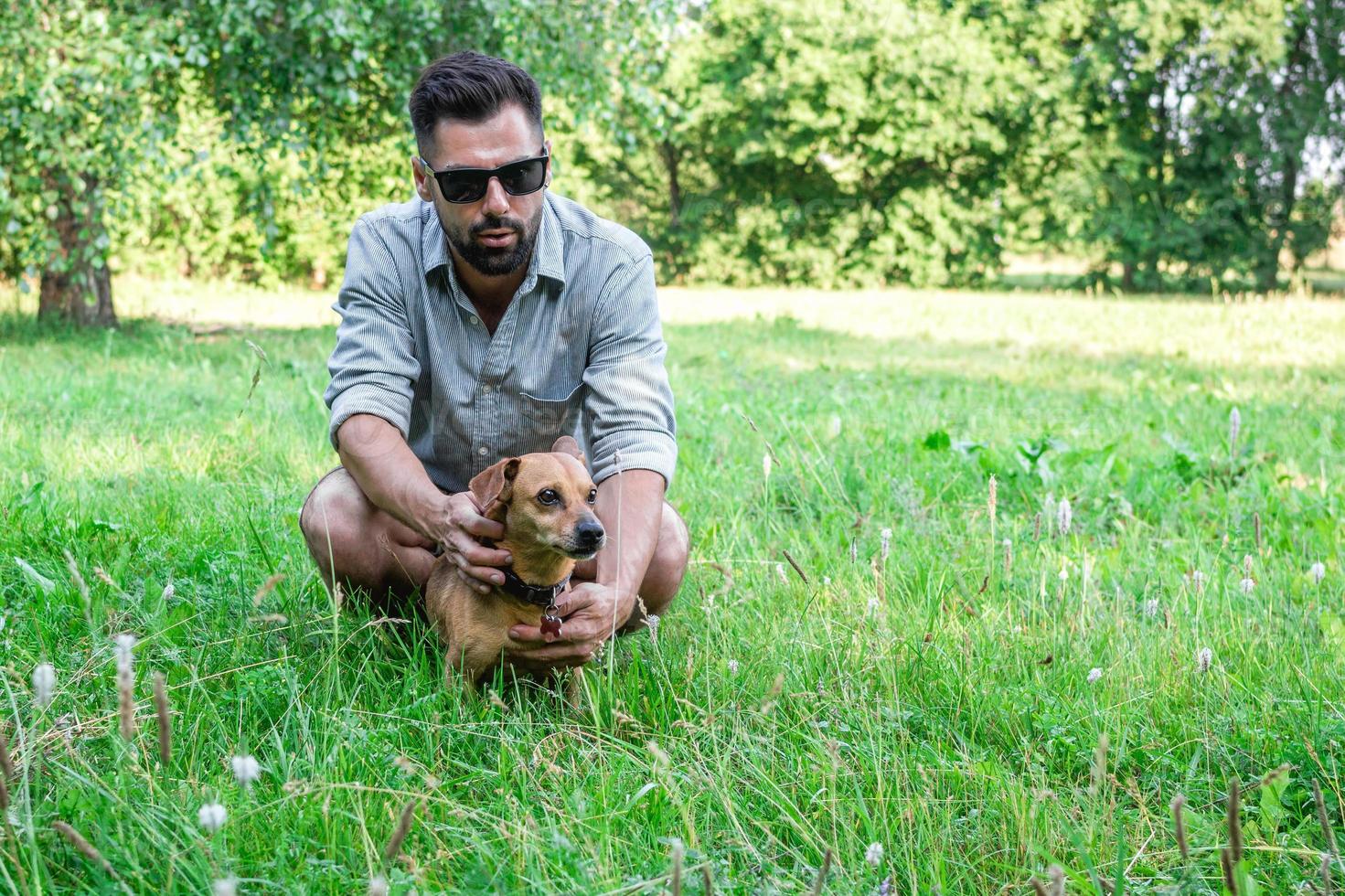 Handsome stylish European man sitting on grass with his dog in park on a walk. Friendship between human and pets. photo