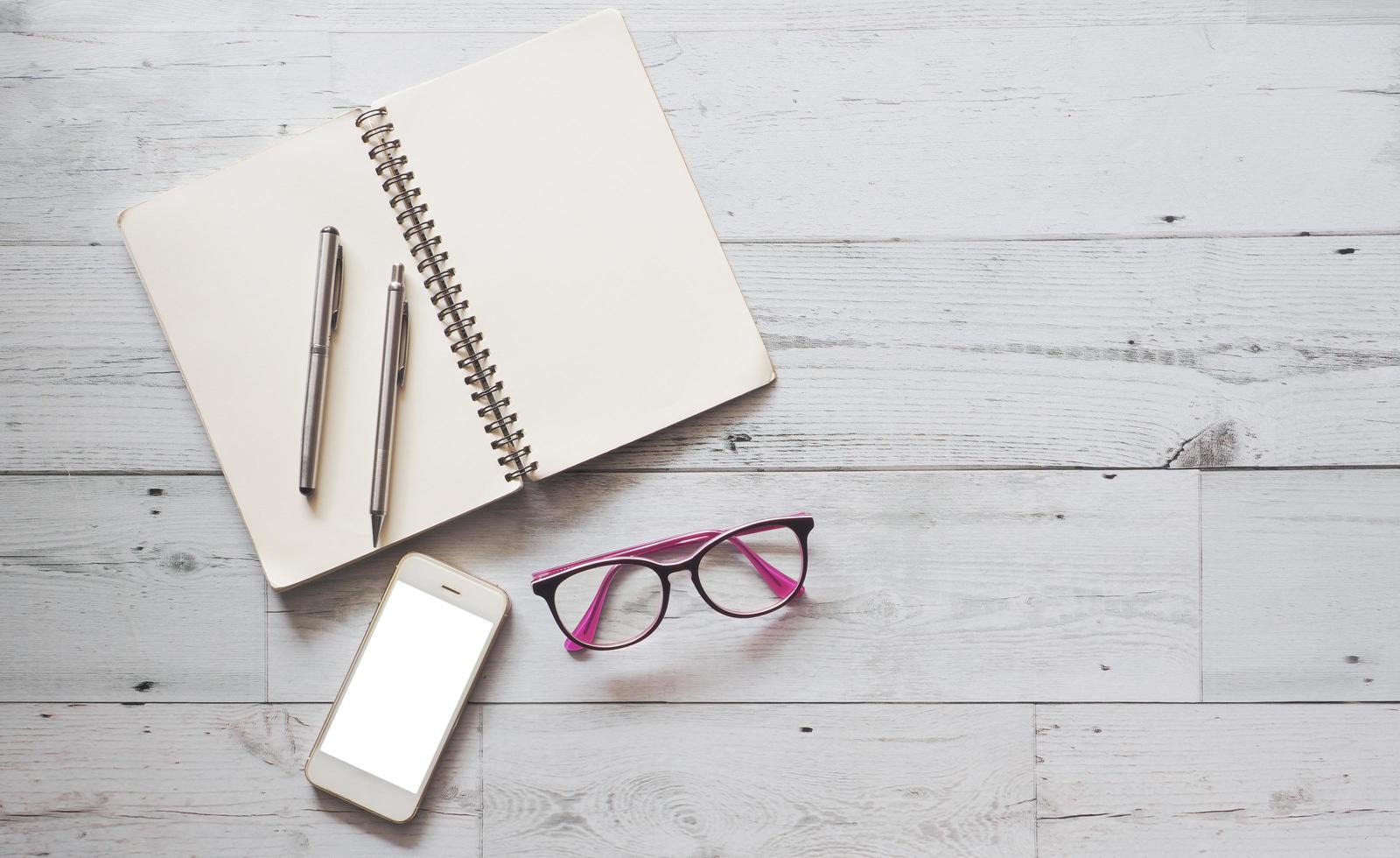 dos bolígrafos plateados en una página en blanco, un cuaderno abierto con un teléfono inteligente y gafas en una mesa de madera blanca, sombra natural y espacio de vista superior claro, concepto de negocio foto