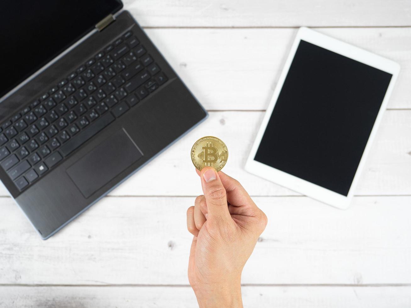 Hand holding gold bitcoin with laptop and tablet on table background top view photo