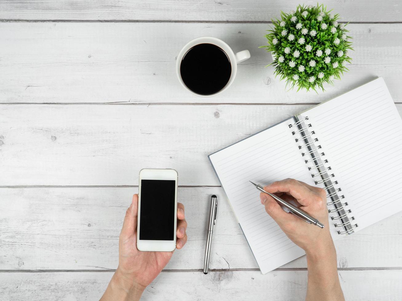 Workspace on white wood table top view copy space,Hand hold silver pen and smartphone in hand with notebook on the desk photo