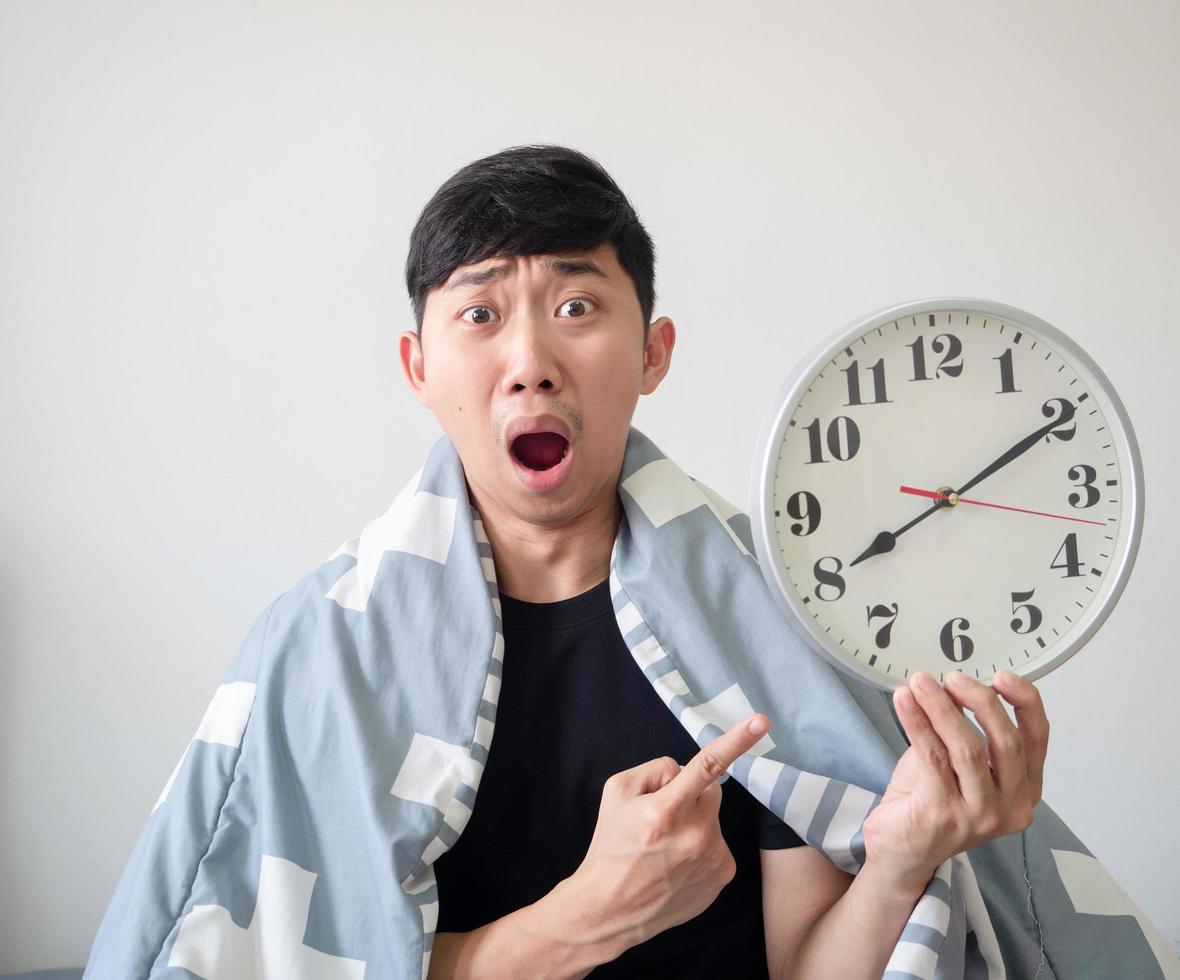 el hombre asiático cubre el punto de la manta en el reloj en la mano con la cara sorprendida concepto tardío fondo blanco foto