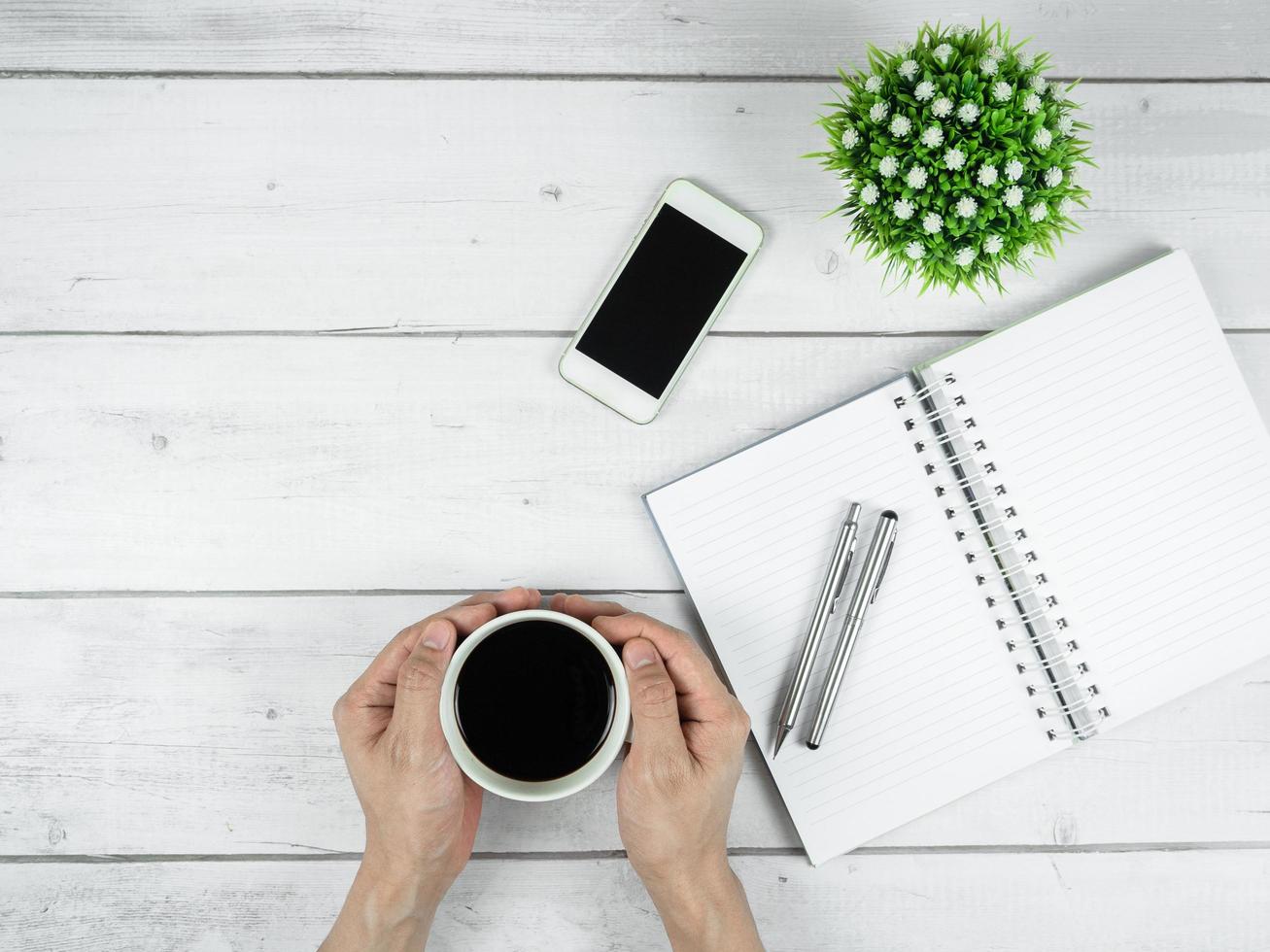 Workspace on white wood table top view copy space,Silver pen on blank page notebook and smartphome black screen with coffee cup in two hand minimal concept photo