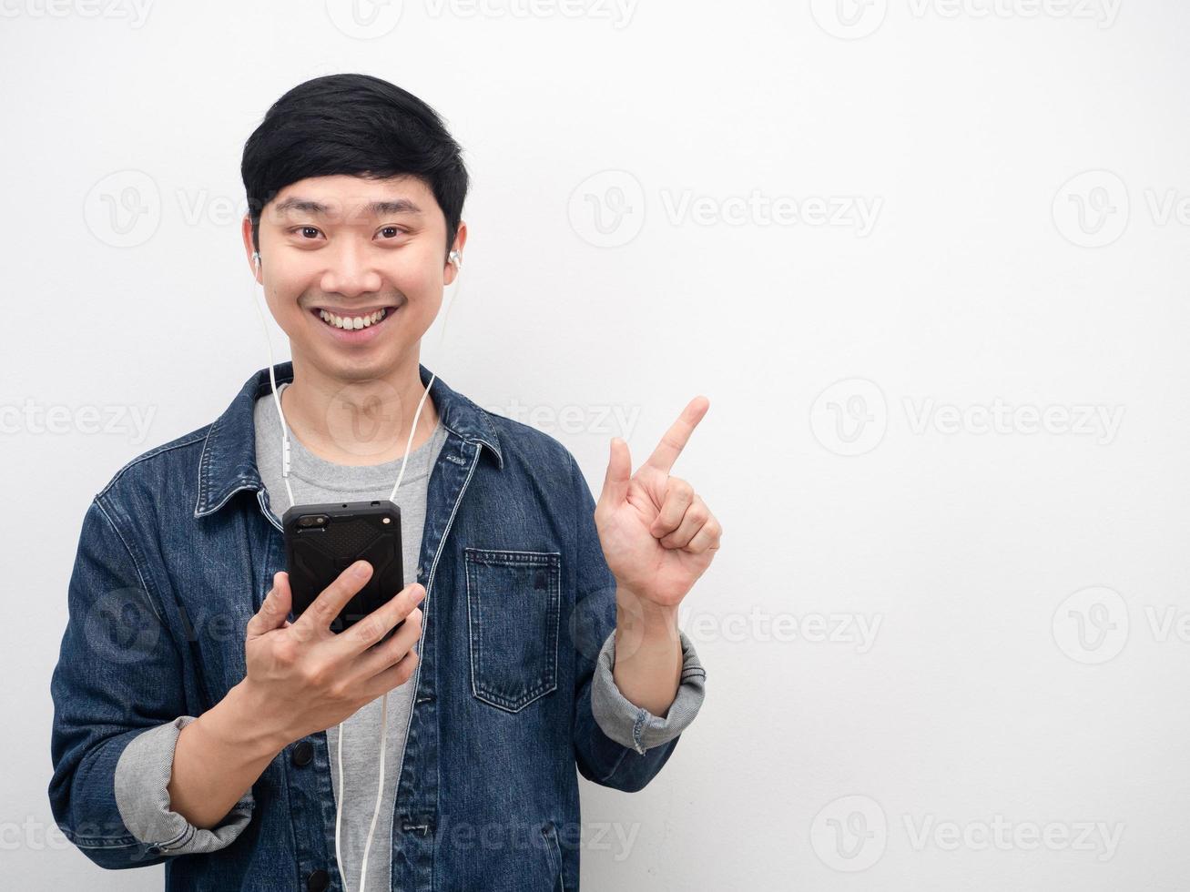 Asian man jeans suit smiling holding mobile phone and using earphone point finger at copy space photo