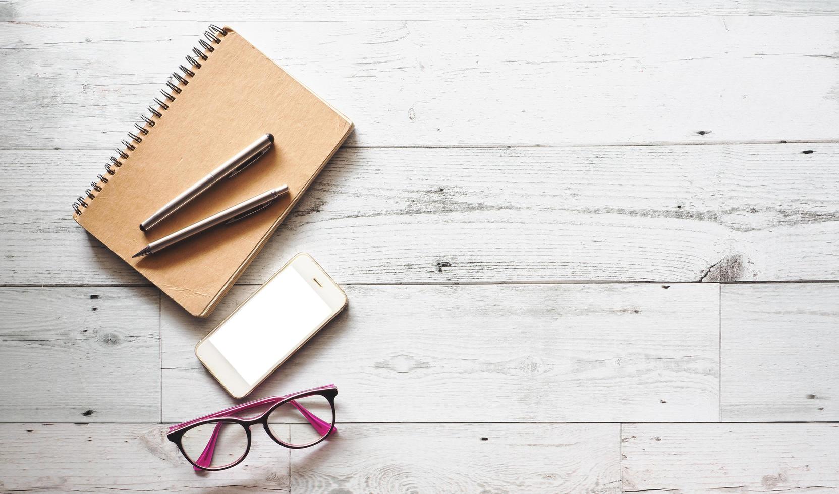 dos bolígrafos plateados en un diario marrón con teléfono inteligente y anteojos en una mesa de madera blanca, sombra natural y espacio de vista superior claro, concepto de negocio foto