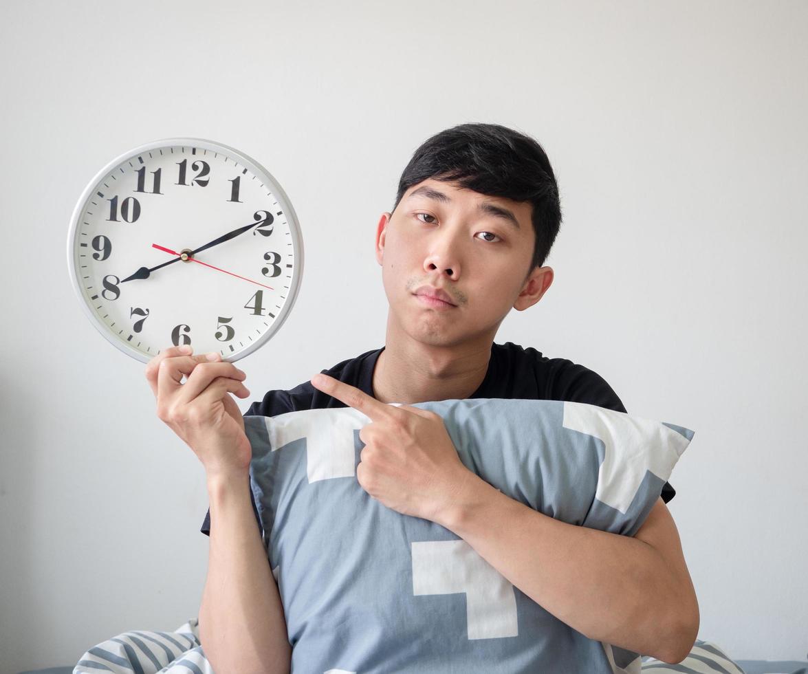 Young man with pillow point finger at clock in hand and feel bored on white isolated work late concept photo