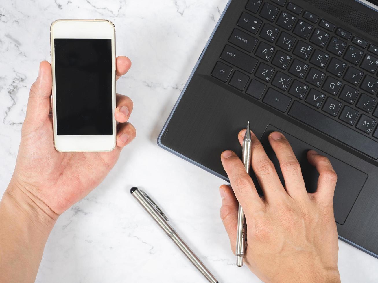 Top view workspace hand holding mobile phone with silver pen and laptop white marble desk photo
