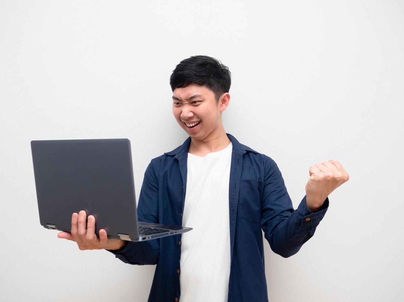 hombre asiático mira la computadora portátil en la mano y siéntete feliz muestra el puño hacia arriba sonrisa feliz en blanco aislado foto