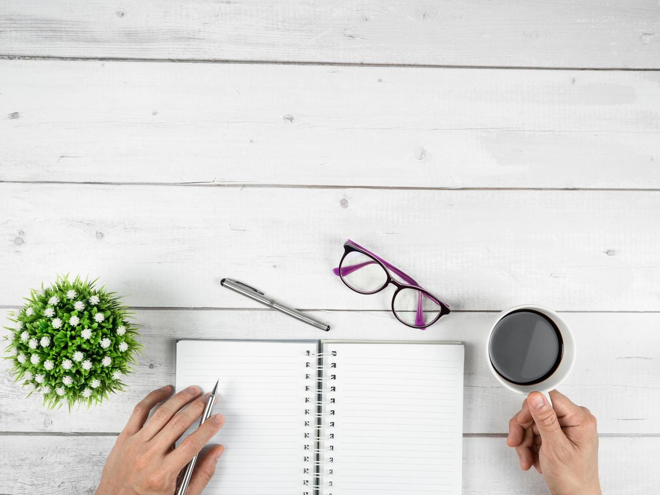 espacio de copia de la vista superior del espacio de trabajo de la oficina, mano con bolígrafo plateado y taza de café y vasos y cuaderno de páginas en blanco sobre una mesa de madera blanca foto