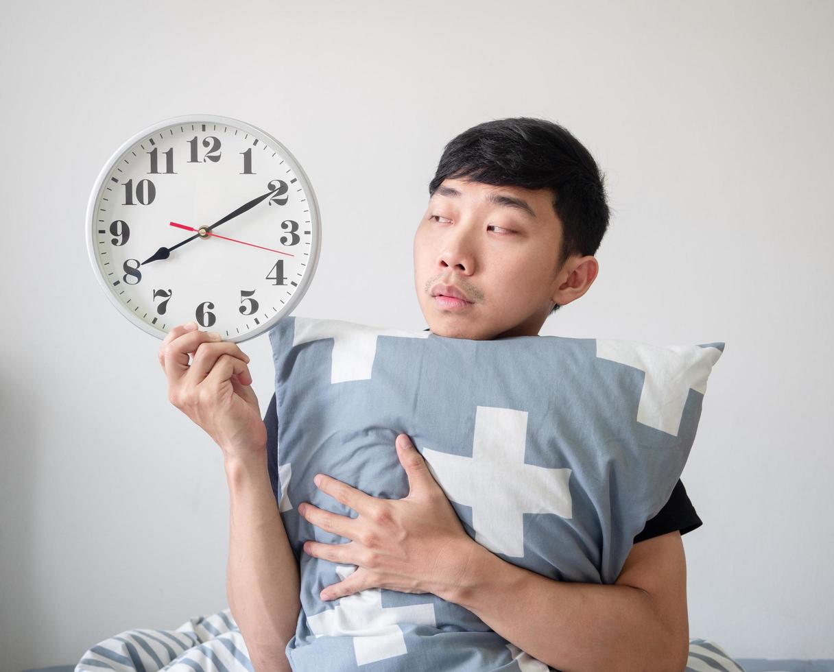 Young man wake up hugging pillow and look at clock in hand feel bored at face on white isolated wake up late concept photo