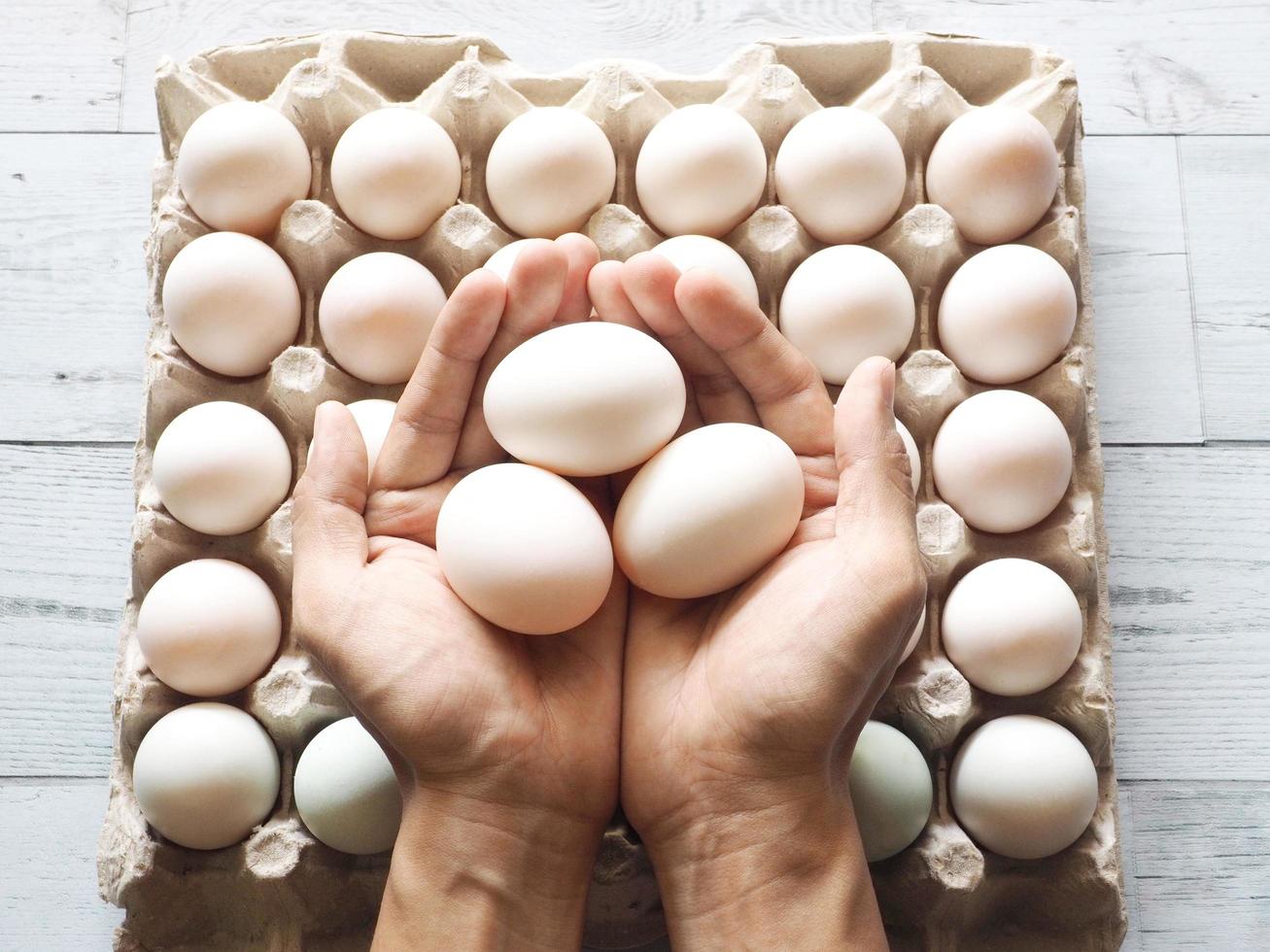 Three duck eggs in hand on group duck eggs in carton box with nature light and shadow on white wood background top view photo