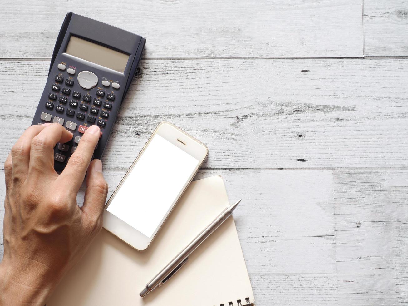 Hand press button of calculator with silver pen smartphone white display and blank page notebook on white wood table nature shadow top view copy space,Business concept photo