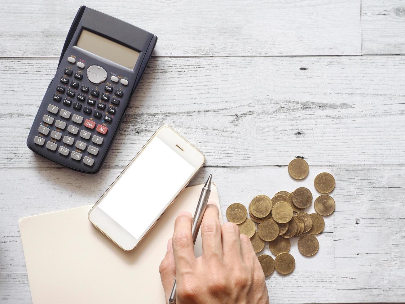 Smartphone white screen and hand holding silver pen on blank page notebook with calculator and gold coins group on white wood table top view above space,Business concept photo