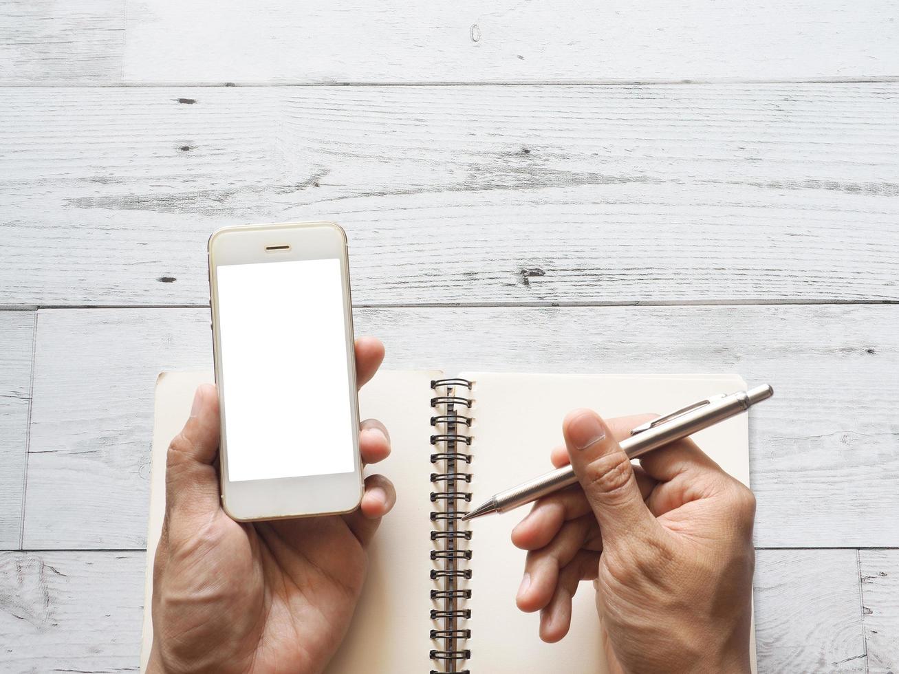 Hand holding smartphone white display and silver pen on blank page notebook on white wooden table top view space nature shadow photo