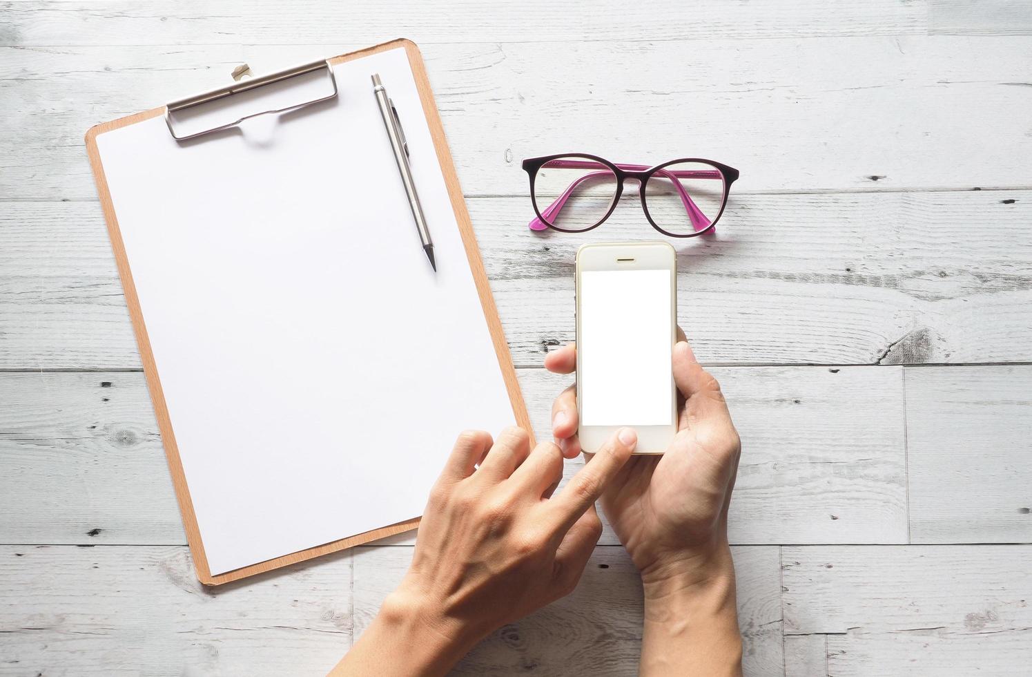 Hand using smartphone white display with silver pen on clipboard and glasses and on white wood table nature shadow top view,Business concept photo